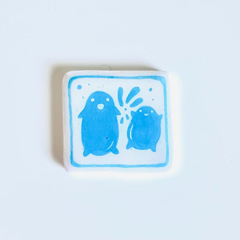 May include: A square, white ceramic tile with a hand-painted design. Two blue, cartoon-like figures are depicted, one larger than the other, with a splash of water or a similar element between them. The figures have simple features and are set against a light blue background.