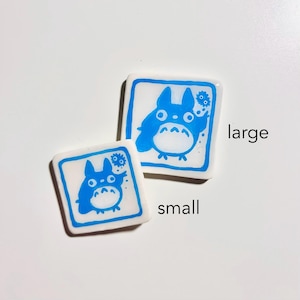 May include: Two square, white ceramic tiles with blue illustrations of a cartoon character. The larger tile has the word "large" next to it, and the smaller tile has the word "small" next to it. The cartoon character has large eyes and a round belly.