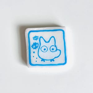 May include: A square, white eraser with a blue border. The eraser features a cartoon illustration of a white creature with large eyes and a small body. Two blue flower-like shapes are on the left side of the creature.