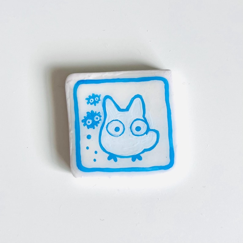 May include: A square, white eraser with a blue border. The eraser features a cartoon illustration of a white creature with large eyes and a small body. Two blue flower-like shapes are on the left side of the creature.