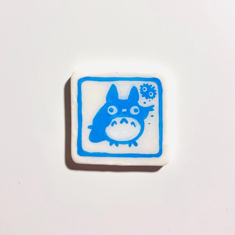 May include: A square, white eraser with a blue border and a blue cartoon character illustration. The character has large eyes, a round belly, and pointed ears. A smaller blue design is in the upper right corner.