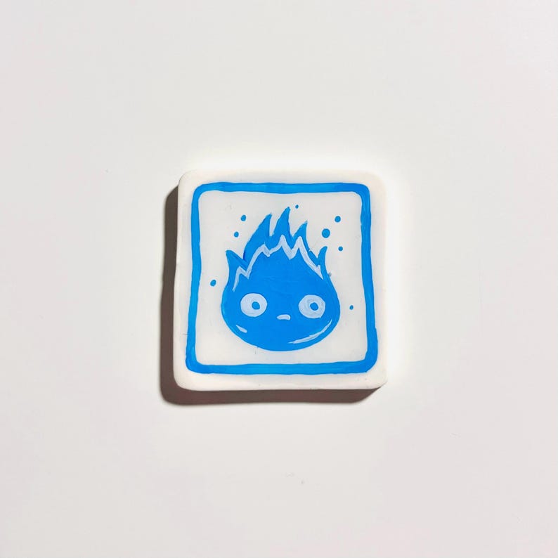 May include: A square, white ceramic tile with a blue cartoon flame design. The flame has a face and is outlined in blue, with a blue border around the tile. Small blue dots surround the flame.