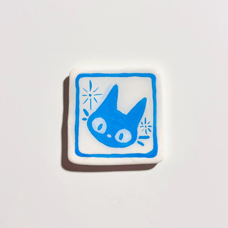 May include: A square, white ceramic tile with a blue cat face design. The cat has large, round eyes and pointed ears, framed by a blue square and starburst accents. The design is hand-painted.
