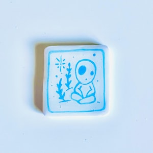 May include: Square, white resin art piece with a blue outline and a blue illustration. The illustration depicts a seated figure with large eyes, surrounded by stylized plants and a starburst design. The art piece is on a white surface.