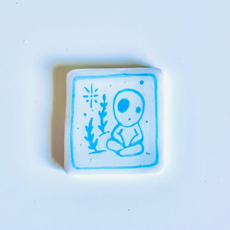 May include: Square, white resin art piece with a blue outline and a blue illustration. The illustration depicts a seated figure with large eyes, surrounded by stylized plants and a starburst design. The art piece is on a white surface.