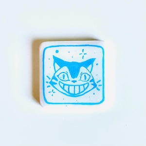 May include: A square, white pin with a blue outline and a smiling cat face design. The cat has a wide grin, large eyes, and pointed ears. The pin is decorated with small star-like accents.