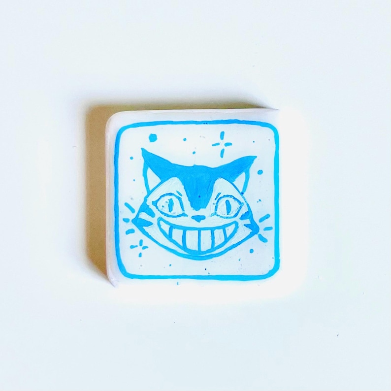 May include: A square, white pin with a blue outline and a smiling cat face design. The cat has a wide grin, large eyes, and pointed ears. The pin is decorated with small star-like accents.