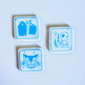 May include: Three square ceramic tiles with blue illustrations on a white background. One tile features two penguin-like figures, another depicts a seated figure, and the last shows a smiling cat face. The tiles have a blue border.