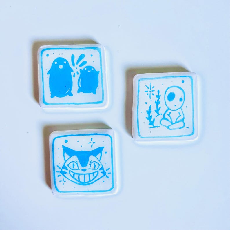 May include: Three square ceramic tiles with blue illustrations on a white background. One tile features two penguin-like figures, another depicts a seated figure, and the last shows a smiling cat face. The tiles have a blue border.