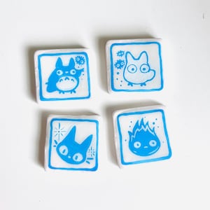 May include: Four square, white-based tiles with blue cartoon character designs. Each tile has a blue border. The characters include a Totoro-like creature, a small, round creature, a cat-like character, and a fire demon.