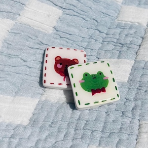 Op de afbeelding: Twee vierkante, witte decoratieve tegels met handgeschilderde dierenontwerpen. Eén heeft een rode beer, de andere een groene kikker met een rode vlinderdas. Beide hebben een gestippelde rand en zijn geplaatst tegen een blauw-witte achtergrond.