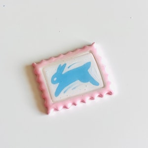 May include: A small, rectangular, pink-framed object with a scalloped edge. The center features a blue rabbit illustration on a white background. The rabbit is depicted in motion, with a sense of movement.