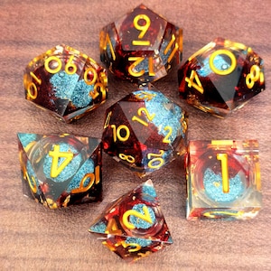 May include: Set of seven polyhedral dice with a red and gold color scheme, with blue glitter accents. The dice have gold numbers and are designed for tabletop gaming. The dice are made of resin.