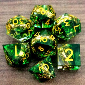May include: A set of seven polyhedral dice with a green and gold color scheme. The dice are transparent with gold-colored numbers and decorative elements. The dice are arranged in a circular pattern, showcasing their various shapes and sizes.