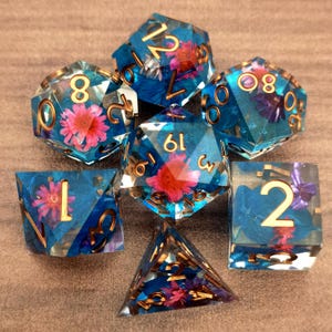 Könnte beinhalten: Ein Satz von sieben polyedrischen Würfeln für Rollenspiele. Jeder Würfel ist transparent mit einem blauen Farbton und enthält rosa und lila Blumen. Goldfarbene Zahlen sind auf jeder Seite sichtbar. Die Würfel liegen auf einer braunen Oberfläche.
