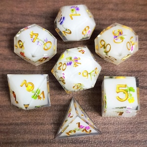 Könnte beinhalten: Set aus sieben polyedrischen Würfeln mit weißer, transluzenter Basis. Jeder Würfel hat goldene Zahlen und zarte Blumendesigns in Lila, Gelb und Rot. Die Würfel sind für Tabletop-Rollenspiele konzipiert.