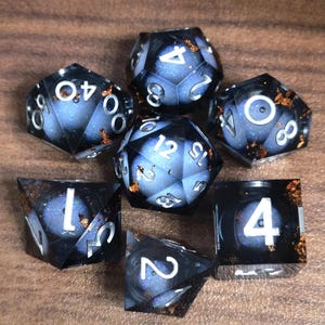 May include: A set of seven polyhedral dice for tabletop games. The dice are black with a blue and glittery interior, and silver numbering. Some dice have gold-colored flakes. The dice are arranged on a wooden surface.