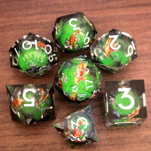 May include: Set of seven polyhedral dice with a koi fish theme. Each die is clear with a green interior, featuring a koi fish and white numbers. The dice are arranged on a brown surface.