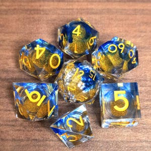 Könnte beinhalten: Sieben polyedrische Würfel mit transparentem Harz, blauen und goldenen Glitzerakzenten und gelben Zahlen. Die Würfel sind für Tischrollenspiele konzipiert.