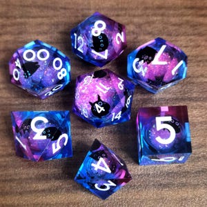 Puede incluir: Un conjunto de siete dados poliédricos con temática de galaxia. Los dados son transparentes con una mezcla de colores azul, morado y rosa. Cada dado presenta números blancos y una silueta de gato negro. Diseñados para juegos de rol.