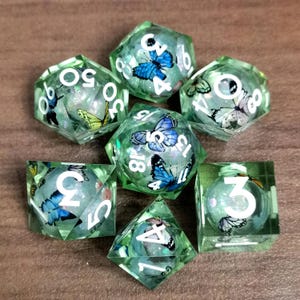 May include: A set of seven polyhedral dice with a green, translucent appearance. Each die has white numbers and is decorated with colourful butterfly inclusions. The dice are designed for tabletop gaming.
