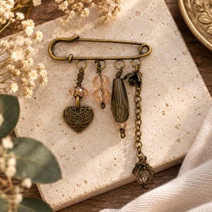 May include: A bronze-toned safety pin brooch featuring four dangling charms. The charms include a filigree heart, two faceted light peach beads, a dark grey teardrop bead, a small metal flower, and a chain ending in a metal flower bead cap.