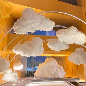 May include: An indoor decorative display featuring multiple fluffy, white cloud-shaped ornaments suspended from curved metal supports. The clouds are illuminated, creating a soft, ethereal ambiance. The background is a warm yellow color.