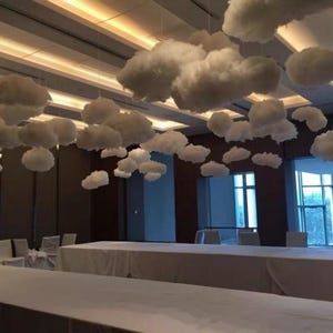 May include: A room decorated with fluffy white cloud decorations suspended from the ceiling. The clouds are of varying sizes and shapes, creating a whimsical atmosphere. Long tables covered in white linens are set up below the clouds.