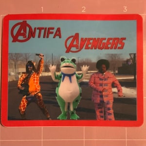 May include: A sticker with a red border featuring the text "Antifa Avengers" above three costumed figures. One is in a giraffe costume, another is a frog, and the third is in a red and white striped suit.