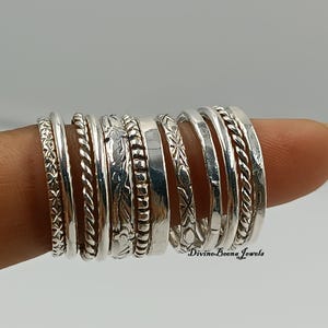 Puede incluir: Una colección de anillos de plata con varios diseños, incluyendo bandas trenzadas, texturizadas y estampadas. Los anillos están apilados, mostrando diferentes estilos y anchos. La imagen está sobre un fondo neutro, resaltando las joyas.
