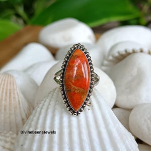 May include: A silver ring featuring an oval, orange gemstone with brown and yellow inclusions. The gemstone is set in a beaded bezel and sits atop a split shank. The ring is displayed on a white seashell.