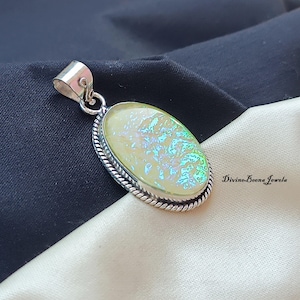 Peut inclure: Pendentif ovale avec une pierre irisée jaune clair sertie dans un cadre en métal argenté. La pierre a une surface texturée avec des reflets bleus et verts. Le cadre présente un motif torsadé. Le pendentif est sur fond noir et crème.