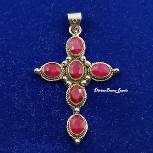 Pode incluir: Um pingente de cruz prateada com pedras preciosas vermelhas ovais vibrantes. O design da cruz apresenta uma argola na parte superior para uma corrente e está sobre um fundo azul escuro. O texto "Divine Beena Jewels" é visível à direita.