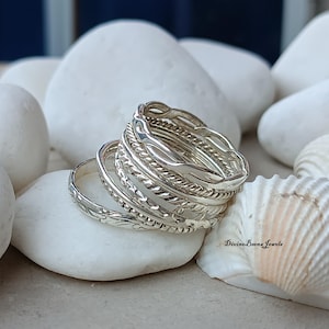 Puede incluir: Un conjunto de anillos de plata con diversas texturas, incluyendo diseños trenzados, con cuentas y estampados. Los anillos se exhiben sobre piedras blancas, con una concha marina en el fondo. Son joyas.