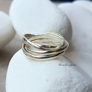 Puede incluir: Un anillo plateado con tres bandas entrelazadas, sobre una piedra blanca. El texto "Divine-Beena Jewels" es visible. El diseño es sencillo y elegante, adecuado para el uso diario.