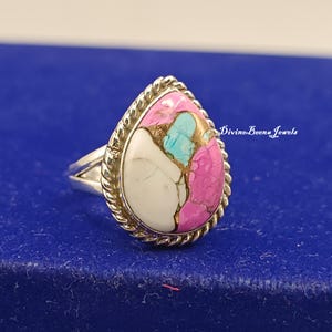 May include: A teardrop-shaped ring with a multi-colored stone. The stone features white, pink, turquoise, and gold hues, set in a silver band with a twisted rope design. The text "Divine Boena Jewels" is visible on the right side.