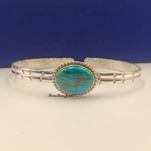 May include: A silver cuff bracelet with an oval, turquoise-colored stone at the center. The stone is surrounded by a twisted rope design. The bracelet has a simple, yet elegant design, with a polished finish. The brand name "Divine Beena Jewels" is printed on the bottom.