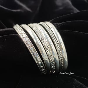 May include: A collection of silver bangles with intricate designs. The bangles feature various patterns, including floral, geometric, and braided textures. The jewelry is displayed on a black surface, highlighting the shine of the metal.