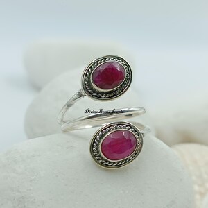 May include: A silver-toned ring featuring two oval-shaped, deep red gemstones. Each gemstone is set within a decorative, textured frame. The ring's design wraps around the finger, with the text "Divine Beena Jewels" visible on the band.