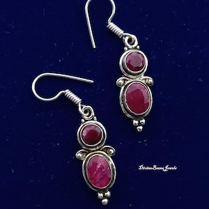 May include: A pair of silver dangle earrings with a dark blue background. Each earring features two red, oval-shaped gemstones and a smaller round gemstone. The earrings have a decorative design and are suspended from a hook.