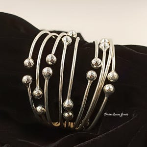 Könnte beinhalten: Ein silberfarbenes Armband mit mehreren gebogenen Stäben, die jeweils in einer kleinen, runden Kugel enden. Das Armband wird vor einem schwarzen Hintergrund präsentiert, der seine reflektierende Oberfläche hervorhebt. Das Schmuckstück hat ein modernes, minimalistisches Design.