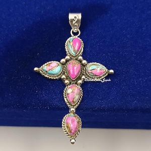 Può includere: Ciondolo a forma di croce argentata con pietre a goccia rosa, turchese e bianche. La croce ha un'ansa decorativa ed è posta su uno sfondo blu scuro. Le pietre sono incorniciate da un motivo a corda intrecciata.
