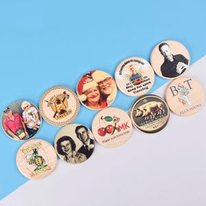 May include: A collection of round wooden coasters with various designs and images. Some coasters feature people, while others display text, logos, and illustrations, including a turtle, cherries, and pigs. The coasters are arranged on a blue and white surface.
