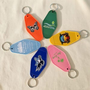 May include: A collection of colorful motel-style keychains. Each keychain features a unique design, including a butterfly, a tree scene, a sunset, and text. The keychains are in various colors, including orange, green, yellow, pink, blue.