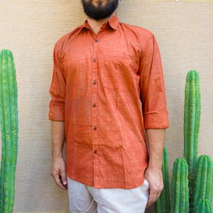 May include: An orange button-down shirt with a chest pocket and long sleeves. The shirt is made of a textured fabric and is paired with off-white pants. The background features green cacti.