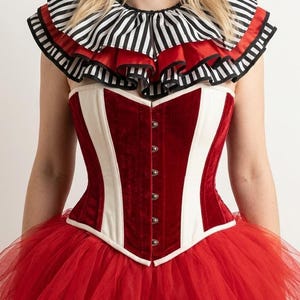 May include: A corset featuring alternating panels of deep red velvet and cream fabric, secured with a front row of silver-toned metal clasps. The garment is paired with a layered, ruffled collar in black and white stripes with red trim.