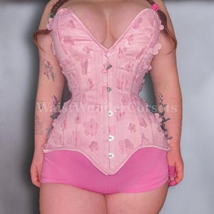 May include: A light pink corset with floral embellishments and a matching pair of shorts. The corset features a sweetheart neckline and a series of metal clasps down the front. The shorts are a solid pink color.