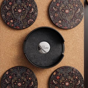 May include: A set of four circular coasters featuring a dark, intricate floral and bird pattern in shades of red, green, and brown. A black circular holder with a central silver-toned disc engraved with the word Flame is included.