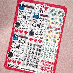 May include: A red sticker sheet with pixelated video game graphics. Includes images of floppy disks, hourglasses, bombs, hearts, and CDs. Text includes "ERROR," "GAME OVER," "NEXT LEVEL," and "Critical error! Insert coffee!"