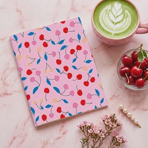 May include: A rectangular notebook with a light pink cover featuring a repeating pattern of red, pink, and peach cherries with blue leaves. Beside it sits a pink mug of green matcha latte, a glass bowl of fresh red cherries, and a pearl hair clip.